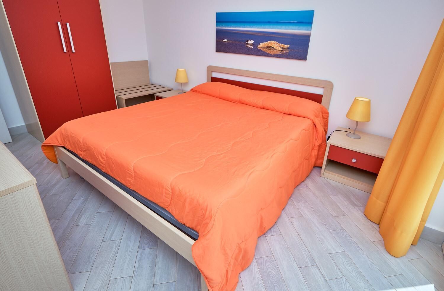 Ein Schlafzimmer mit einem Bett, einem Nachttisch, einer Lampe, einem roten Kleiderschrank und einem Bild von einer Strandlandschaft.