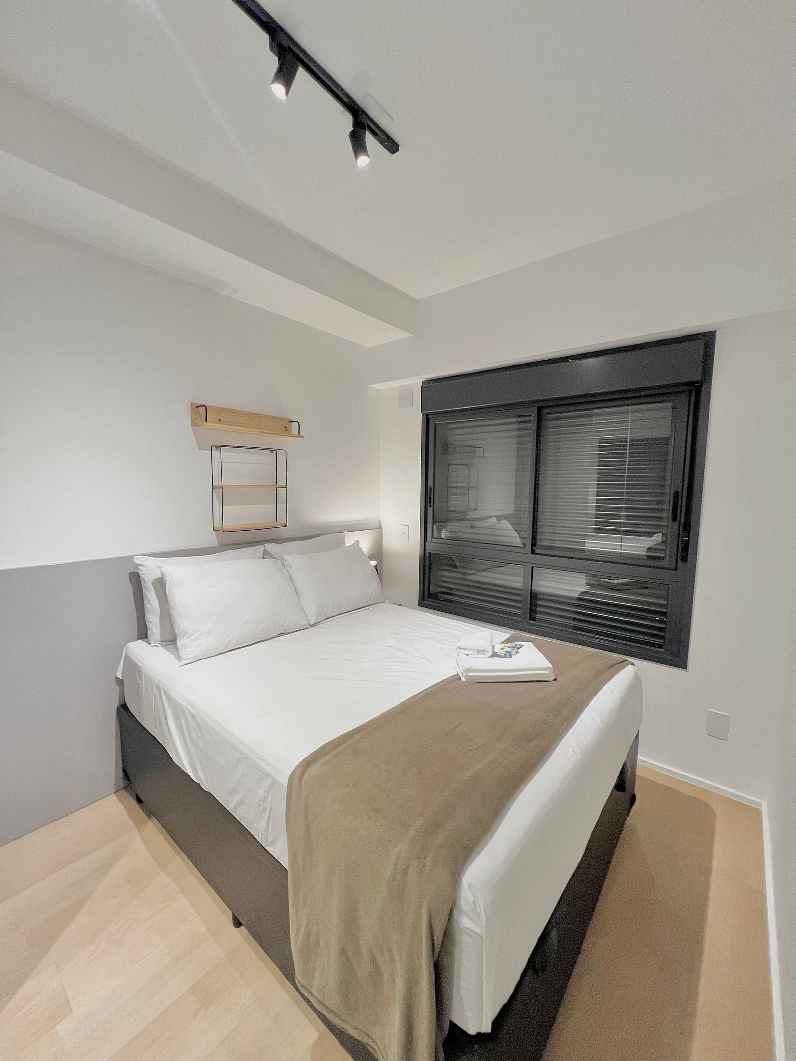 A modern bedroom with a bed, a window with blinds, a bedside table, and a ceiling light fixture.