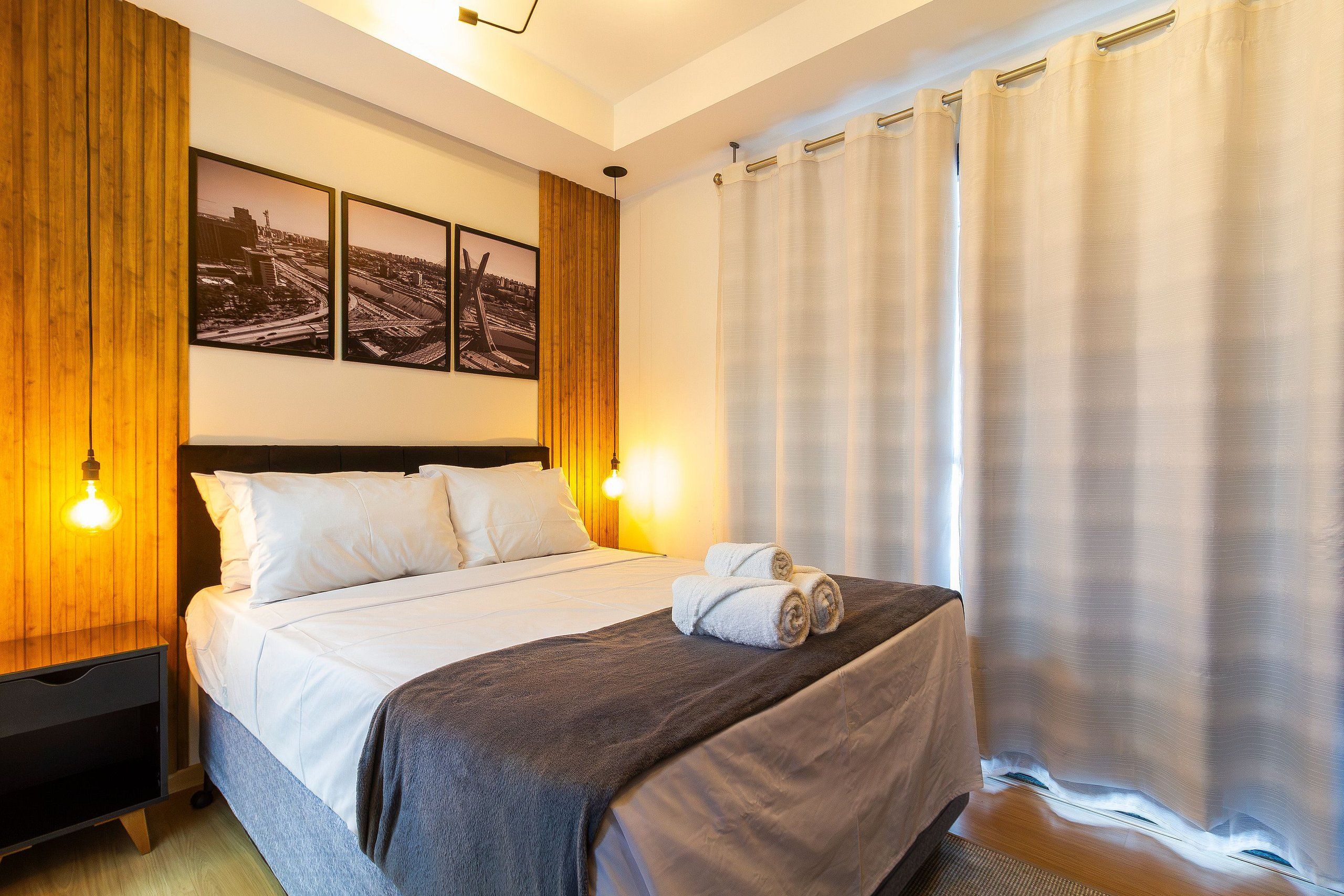 A hotel room with a bed, two wall-mounted lamps, a large window with curtains, and two framed pictures on the wall.