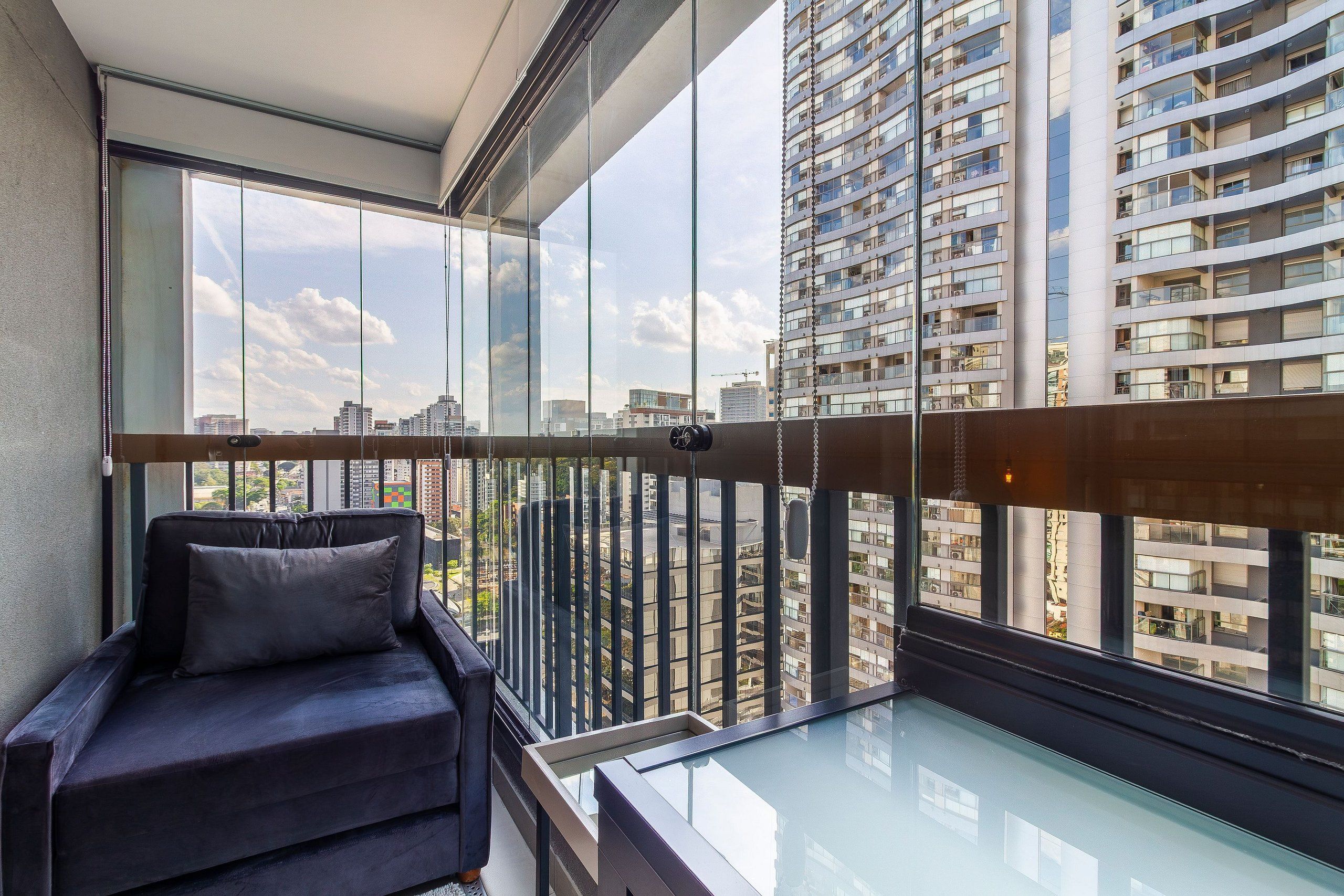 Un balcón moderno con barandilla de vidrio, un sofá de color oscuro y vistas a edificios altos.