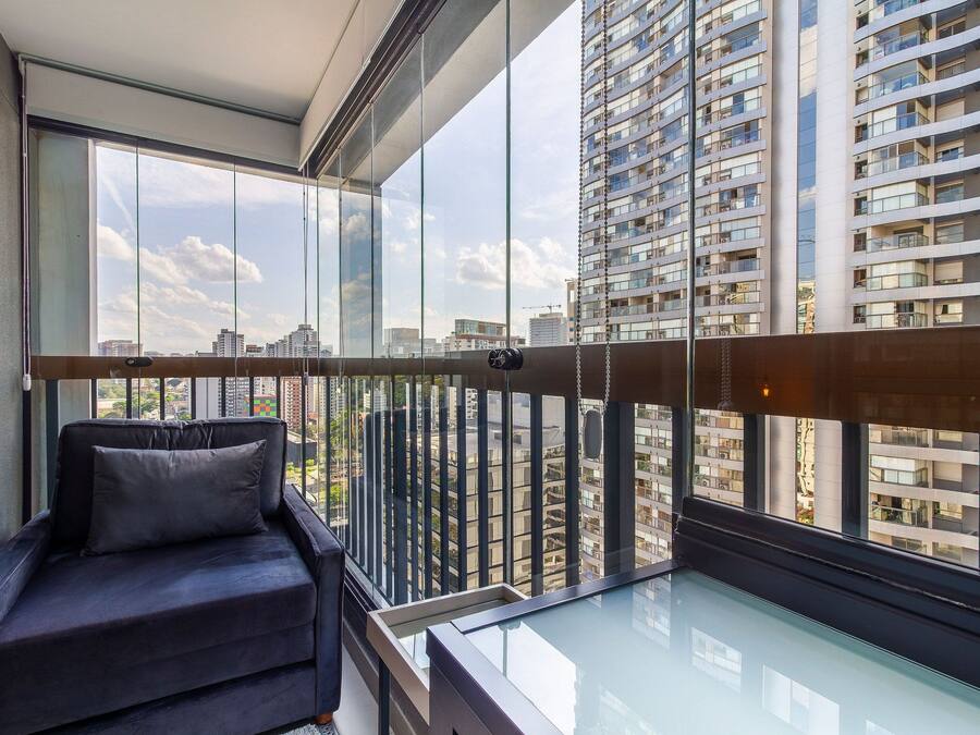 En moderne balkon med gelænder i glas, en mørk sofa og udsigt til høje bygninger.