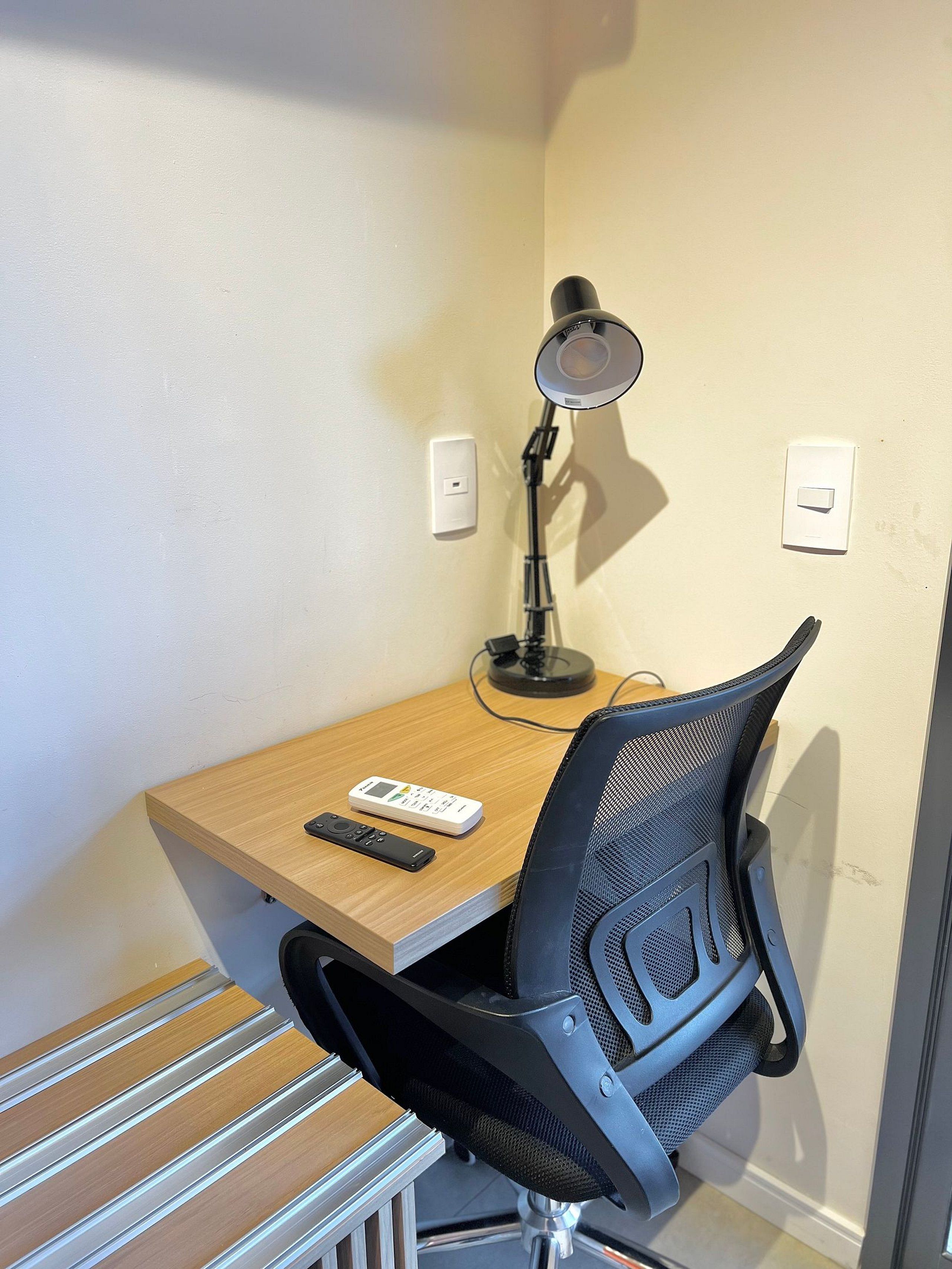 A corner desk with a chair, a desk lamp, a remote control, and a power strip.