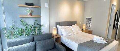 A modern bedroom with a grey sofa, a bed with white bedding, a bedside table with a lamp, and wall-mounted shelves with plants.