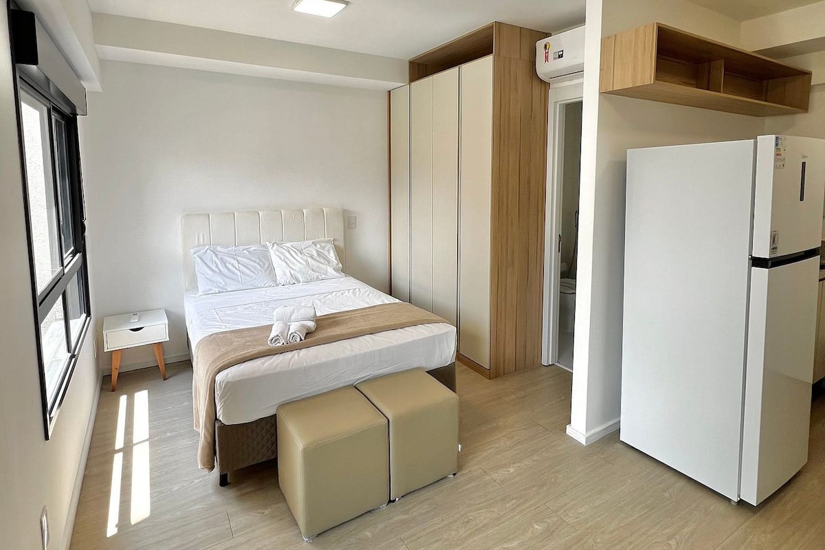 Un dormitorio con cama, mesitas de noche, un banco, un armario y un refrigerador blanco.