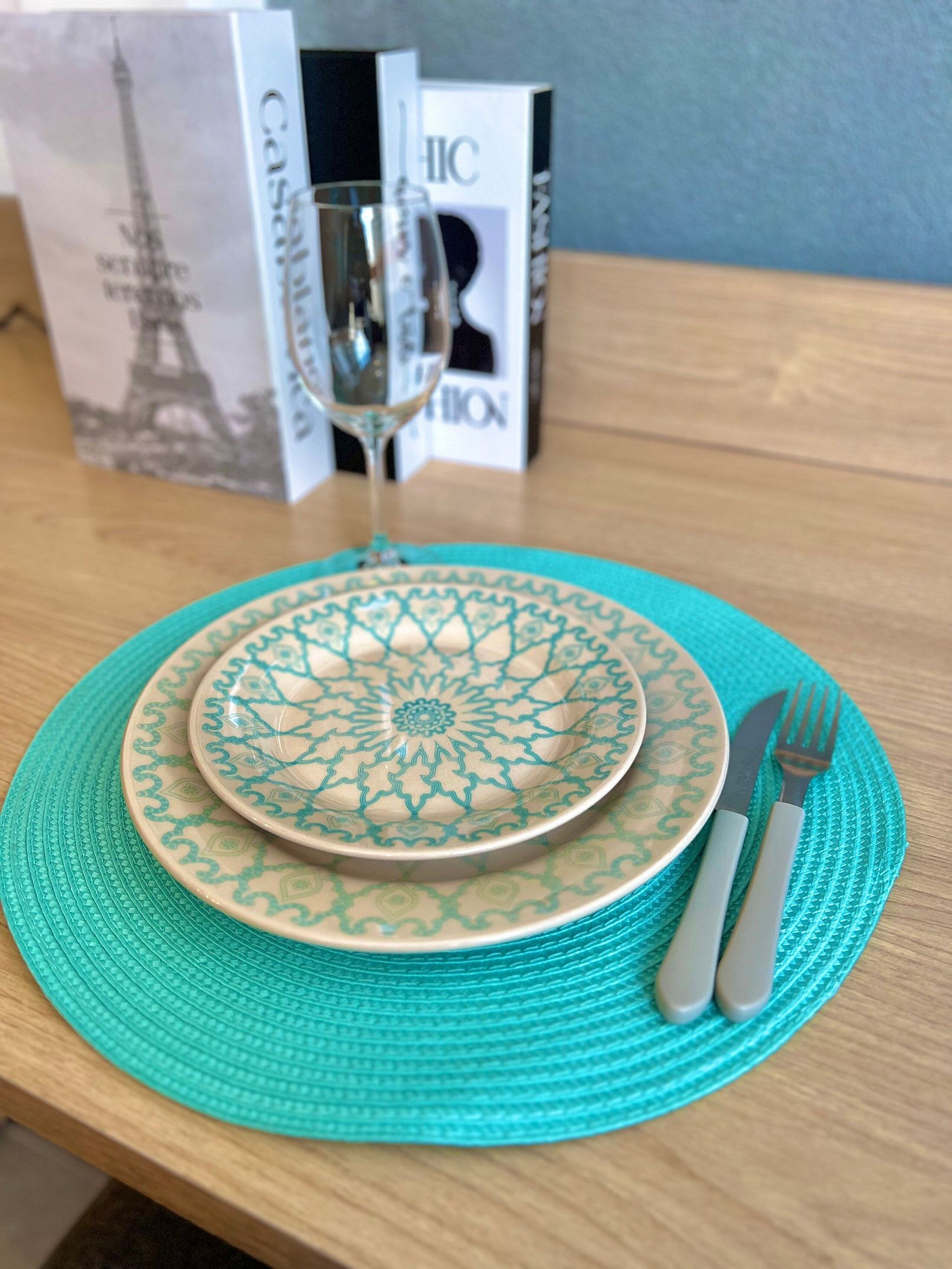 A table with a decorative plate, a glass of water, and a fork and knife on a teal placemat.