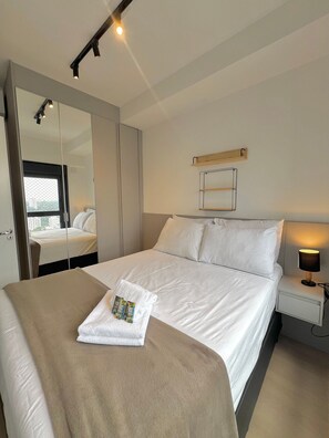 A hotel room with a large bed, a bedside table with a lamp, a mirror, and a window with a view of buildings. - Innvista Uwin Brooklin (São Paulo)