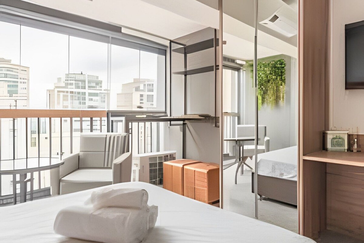 Een moderne hotelkamer met een groot bed, een zithoek met een stoel, een tafeltje, een spiegel en uitzicht op de stad door grote ramen.