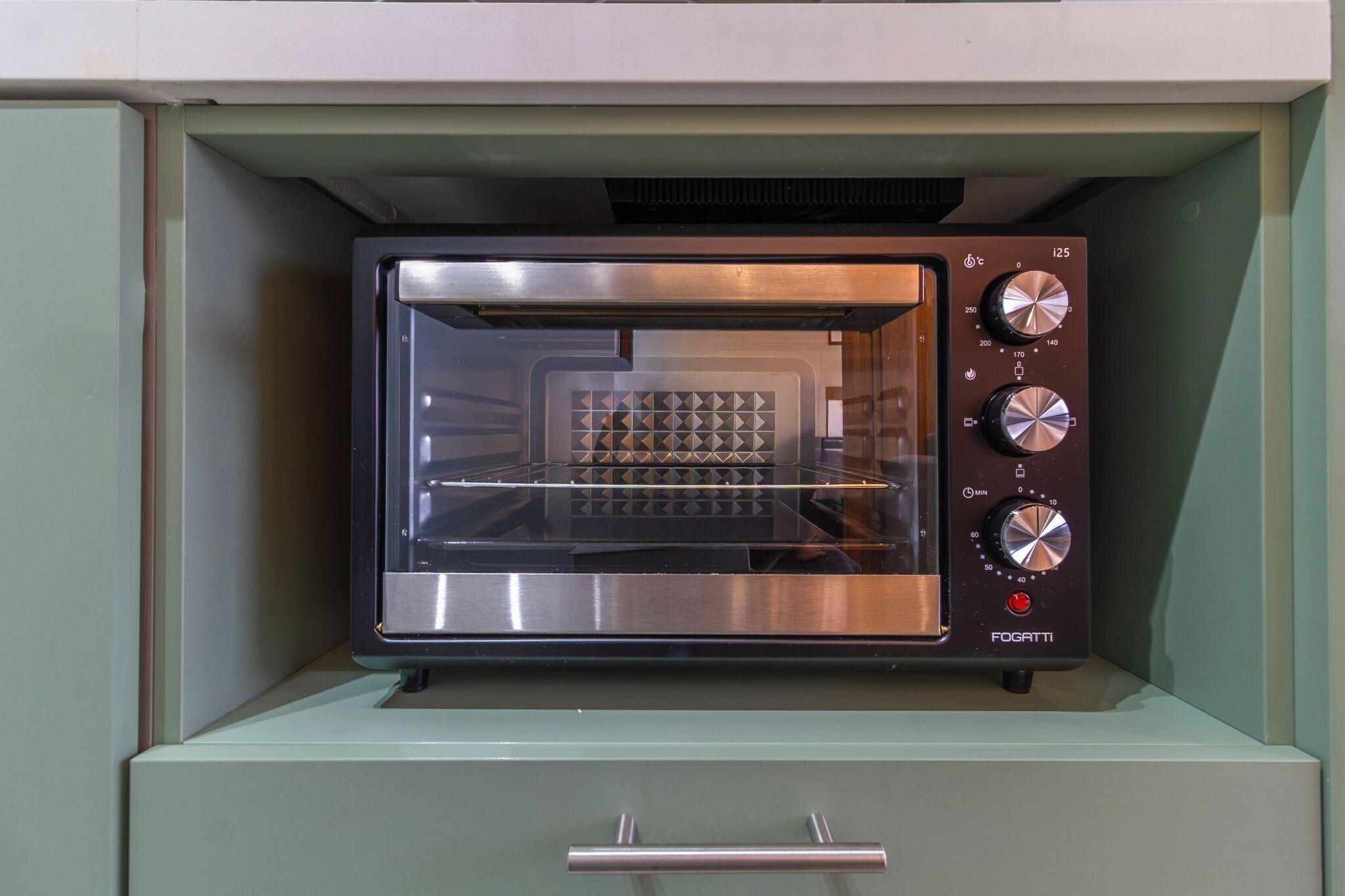 A compact, stainless steel toaster oven with a digital control panel, housed in a green cabinet with a drawer below.