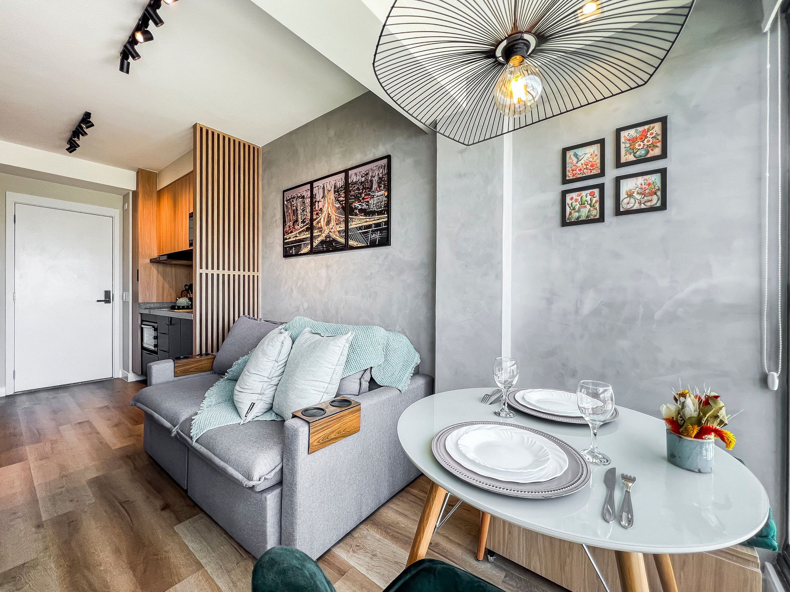 A modern living room with a grey sofa, a dining table set for two, and a kitchen area with wooden cabinets.