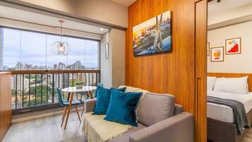 Quarto de hotel moderno com varanda, cama, sofá com almofadas azuis e vista para a cidade.