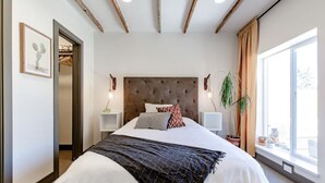 Un dormitorio con una cama grande, techo de madera, mesitas de noche con lámparas, una ventana con cortinas y una planta.
