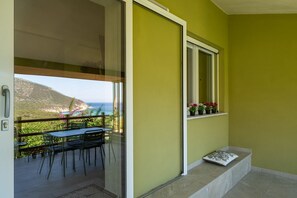 Ein Balkon mit Tisch und Stühlen, ein Fenster mit Blumentöpfen und Blick auf Meer und Berge.