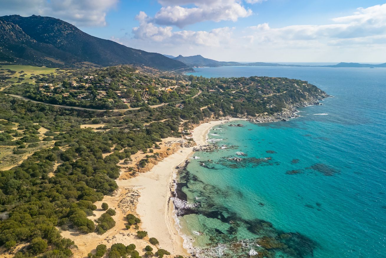 Luftaufnahme einer Küstenlandschaft mit Strand, klarem türkisblauem Wasser und einem gebirgigen Hintergrund.