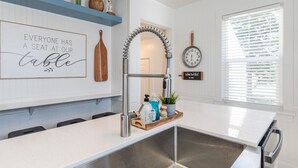 Una cucina moderna con un piano di lavoro bianco, lavello in acciaio inox e uno specchio decorativo. Si vede un orologio appeso al muro e una finestra con persiane.