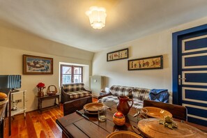 A cozy living room with a sofa, a dining table set with plates, glasses, and a pitcher. There is a television on a stand, a window, and a door with a blue frame.