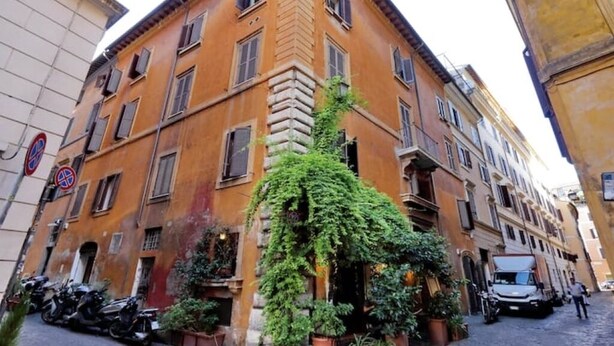 A narrow street with a building featuring a corner with a large, leafy tree. There are parked motorcycles and a truck on the street. - Luxury & Charm between Pantheon and Spanish Steps (Roma)