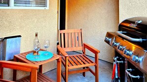 Une terrasse avec une table en bois, deux chaises, une bouteille de vin et un barbecue.