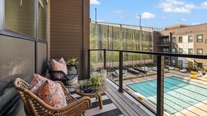 Un balcón con mobiliario de mimbre, una pequeña mesa con plantas y vistas a una piscina y edificios cercanos.