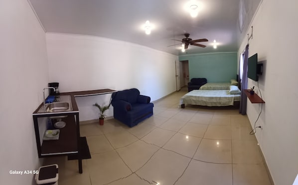 A compact hotel room with a kitchenette, a blue armchair, a bed, a ceiling fan, and a TV mounted on the wall.