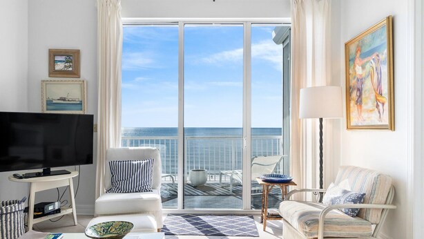 A living room with a large TV, two white sofas, a dining table, and a view of the ocean through large glass doors. - Spanish Key 703 (Pensacola)