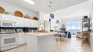 A modern kitchen with white cabinetry, a central island, and a dining area with a view. - Spanish Key 703 (Pensacola)