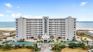 A multi-story hotel with a central entrance, surrounded by a parking area and palm trees, overlooking a beach. - Spanish Key 703 (Pensacola)