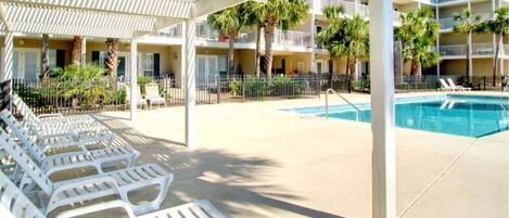 A poolside area with lounge chairs, a pool, and a covered seating area with palm trees.