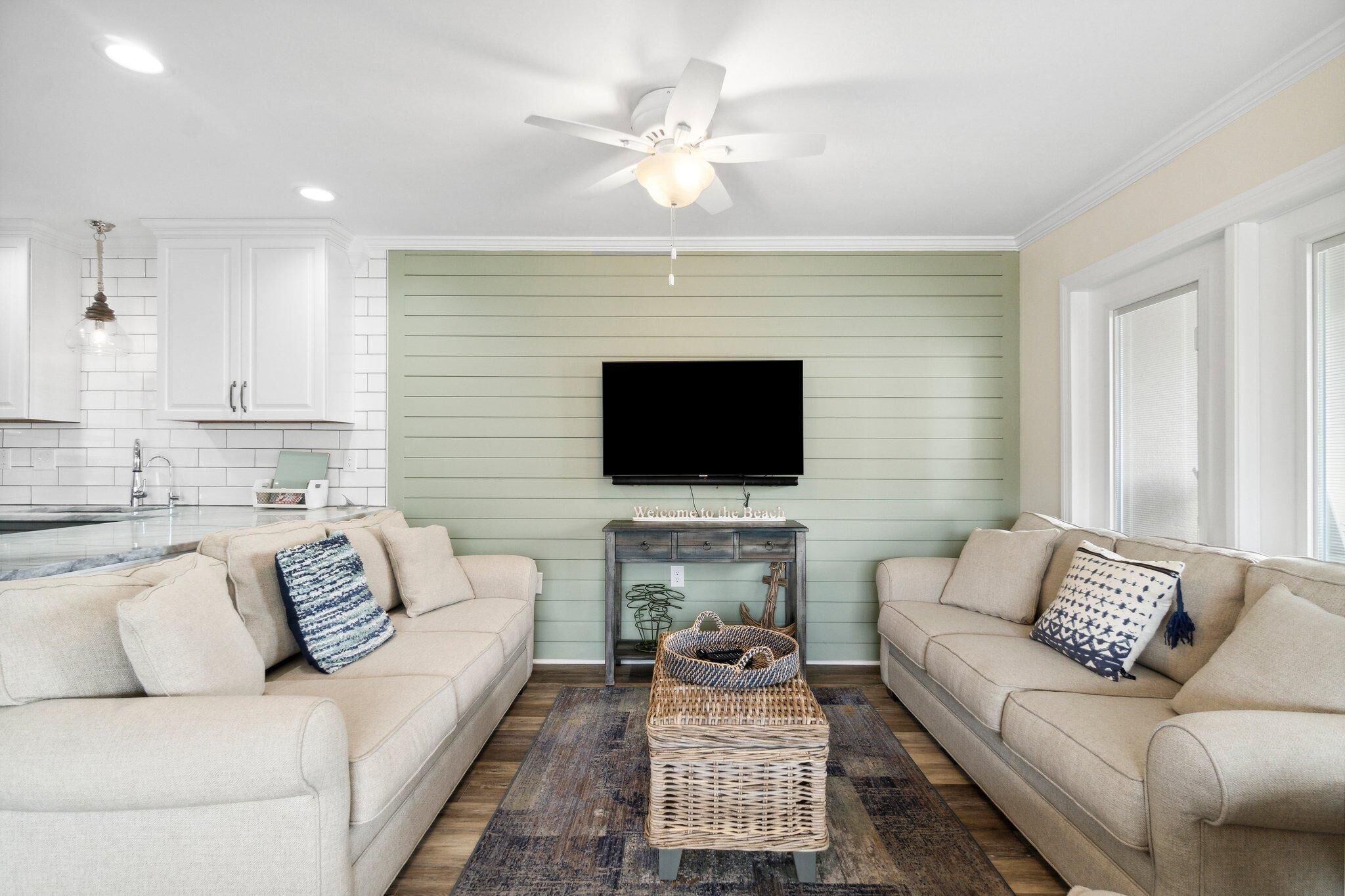A living room with two beige sofas, a coffee table, a television on the wall, and a ceiling fan.
