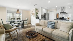 A modern living room with a sofa, a dining table, and a kitchen area with white cabinets and a stainless steel range hood. - Grand Caribbean East 417 (Pensacola)