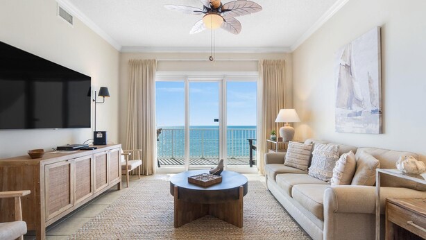 A living room with a large TV, a ceiling fan, a sofa, a coffee table, and a view of the ocean. - Windemere 502 (Pensacola)