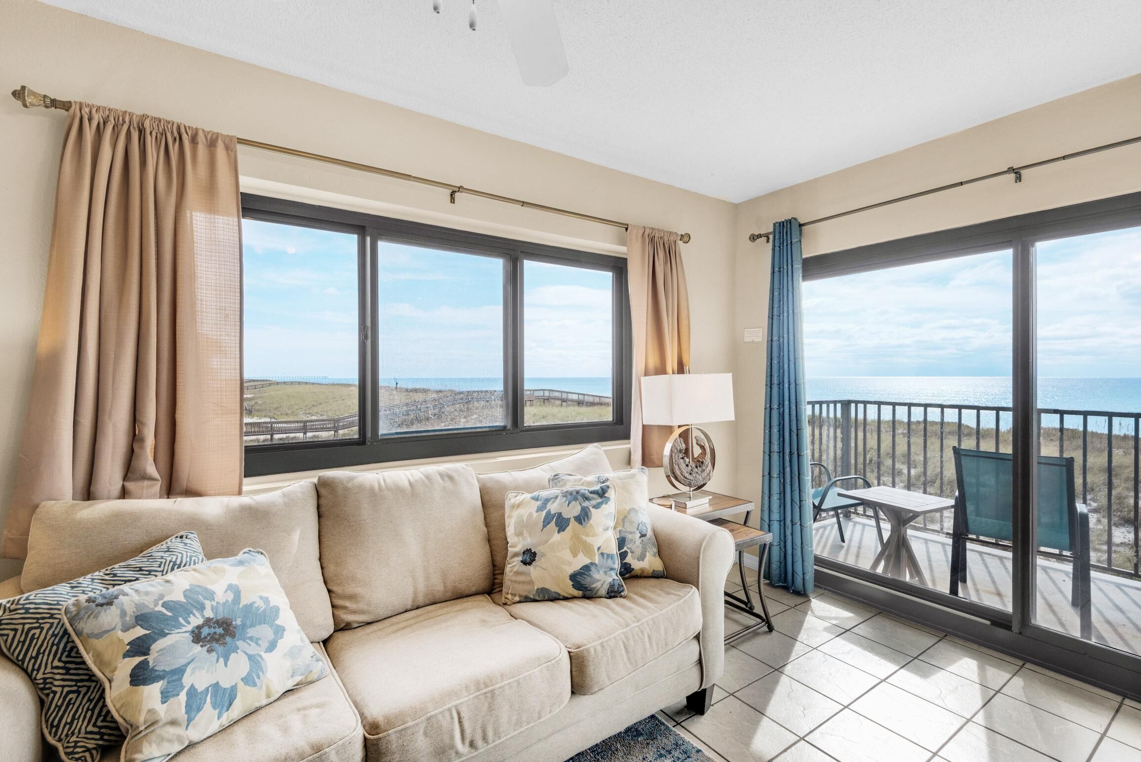 A living room with a beige sofa, floral cushions, a side table with a lamp, and a view of the sea through large windows.