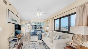 Sala de estar com sofá branco, poltrona azul, mesa de centro e televisão sobre um suporte de madeira. Cozinha ao fundo, com armários azuis e uma janela com cortinas.
