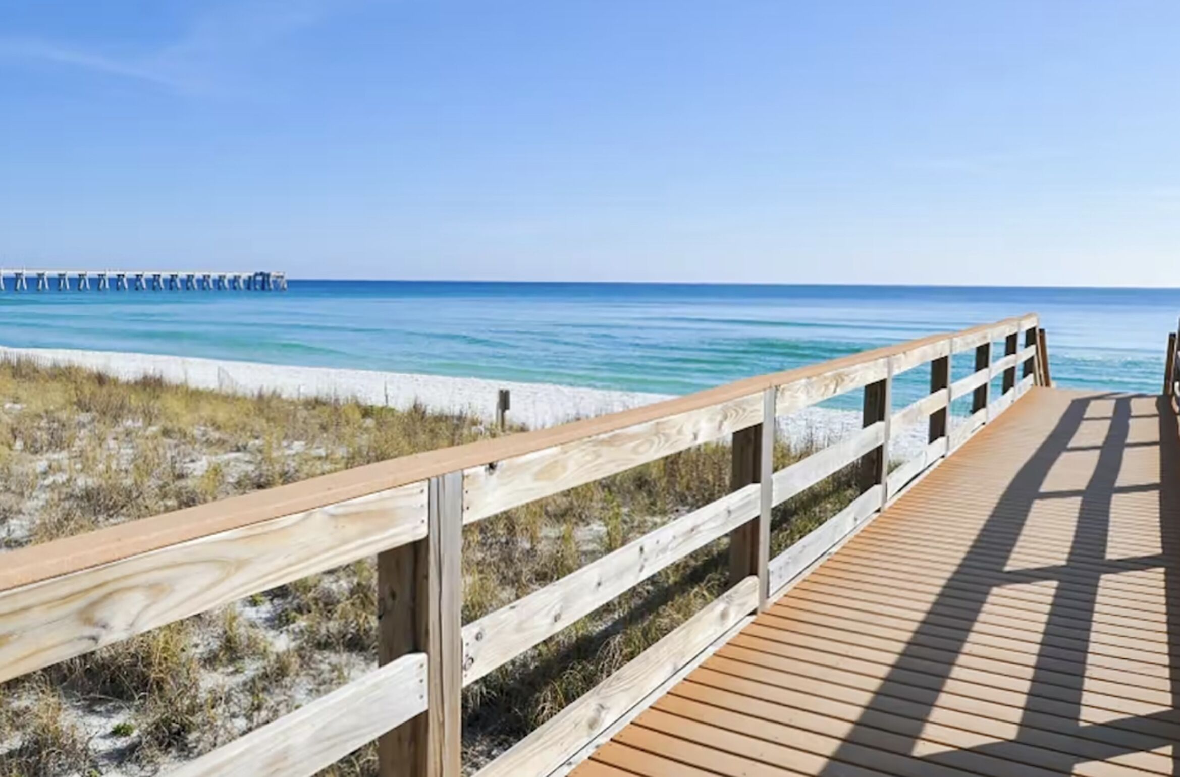 A wooden boardwalk leading to a beach with clear blue water and a pier in the distance.