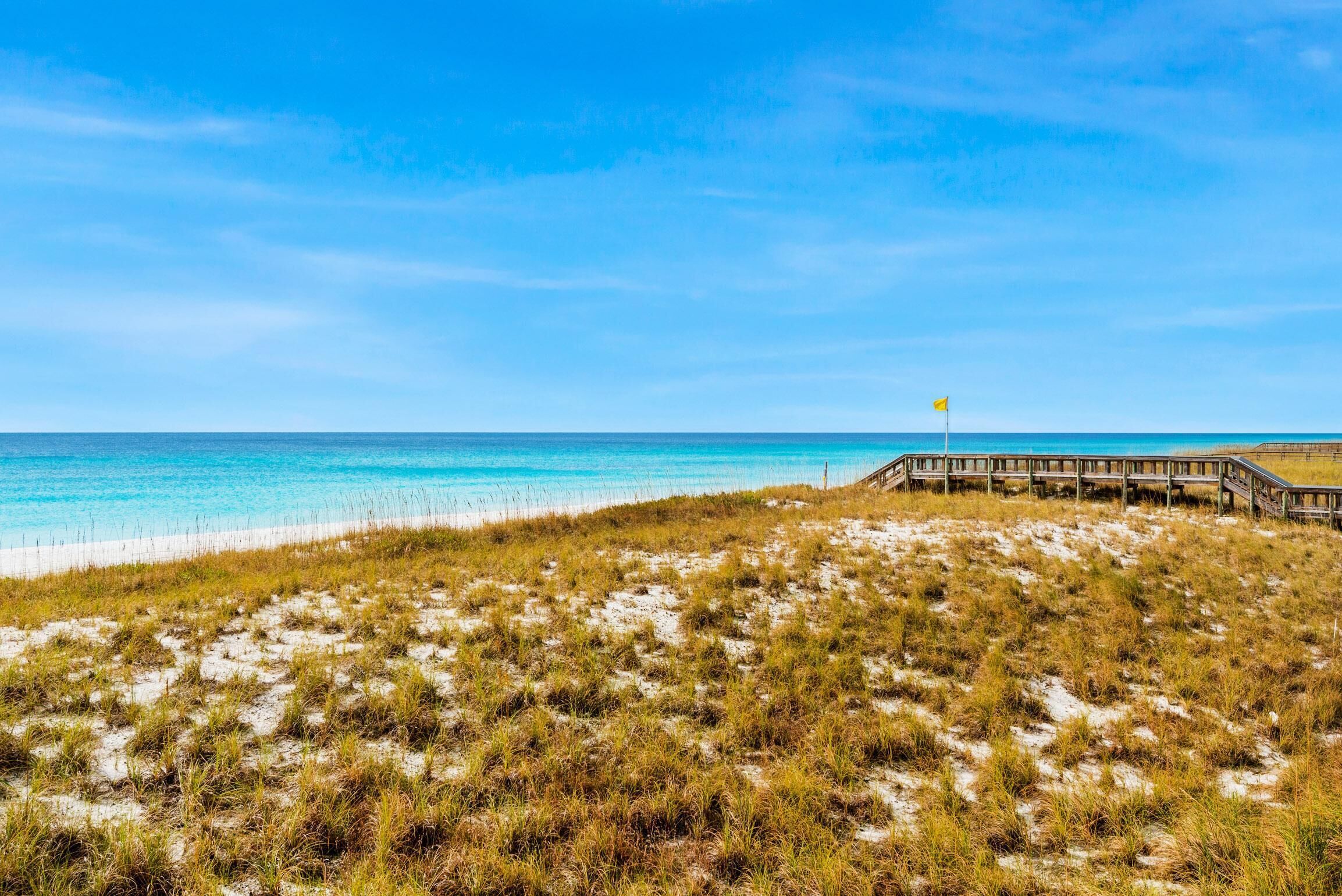 Ein Strand mit einer Holzpromenade, die zu einer Flagge führt, sandigen Dünen und klarem, blauem Wasser.