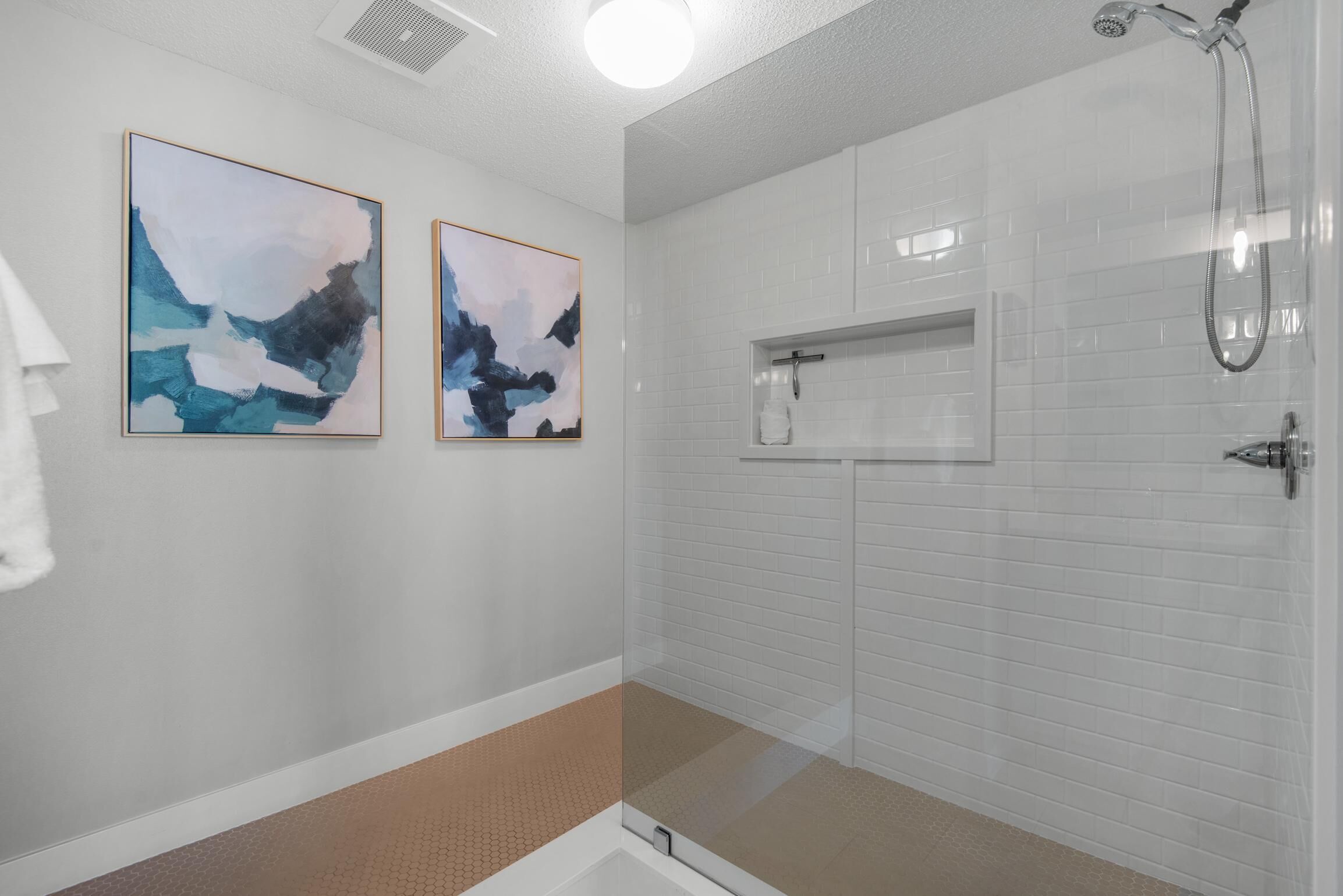 A modern bathroom with white tiled walls, a glass shower enclosure, and two abstract paintings.