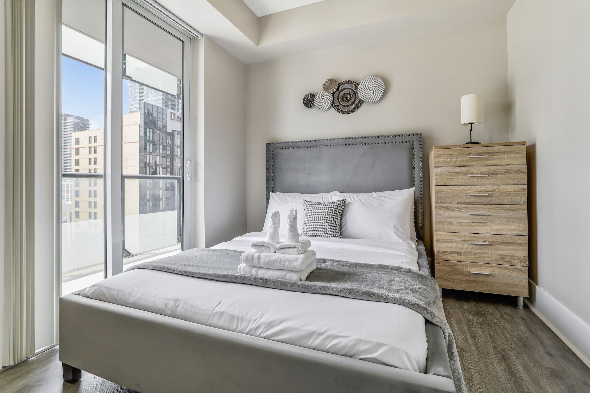Un dormitorio moderno con un ventanal grande, un cabecero tapizado gris, un aparador de madera y un elemento decorativo en la pared.
