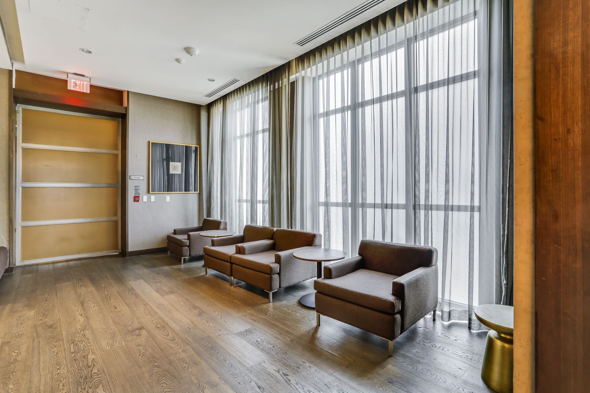 A hotel room with a wooden floor, large windows with sheer curtains, a sofa set, and a small table.