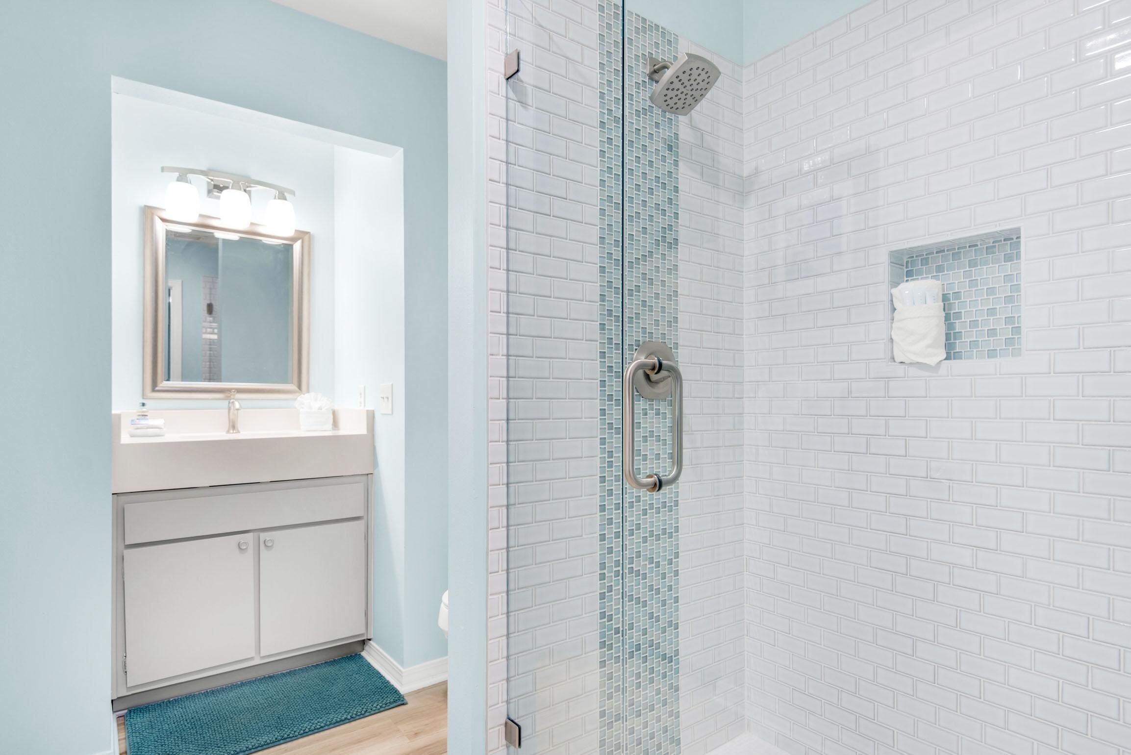 Un baño con ducha envuelta en vidrio, un lavabo blanco con espejo y un tapete azul.