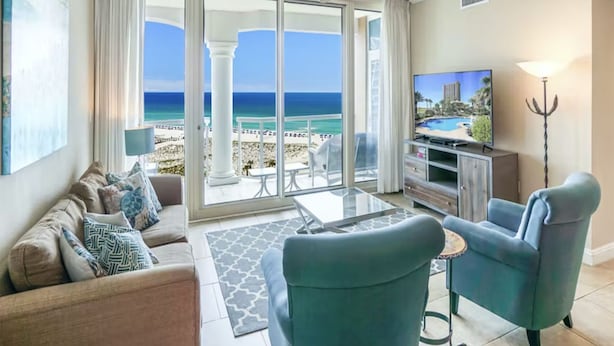 A spacious living room with a large sliding glass door leading to a balcony with a view of the ocean. There is a television on a wooden cabinet, a ceiling fan, and a small table with chairs. - Portofino Island Resort 1- 809 (Pensacola Beach)
