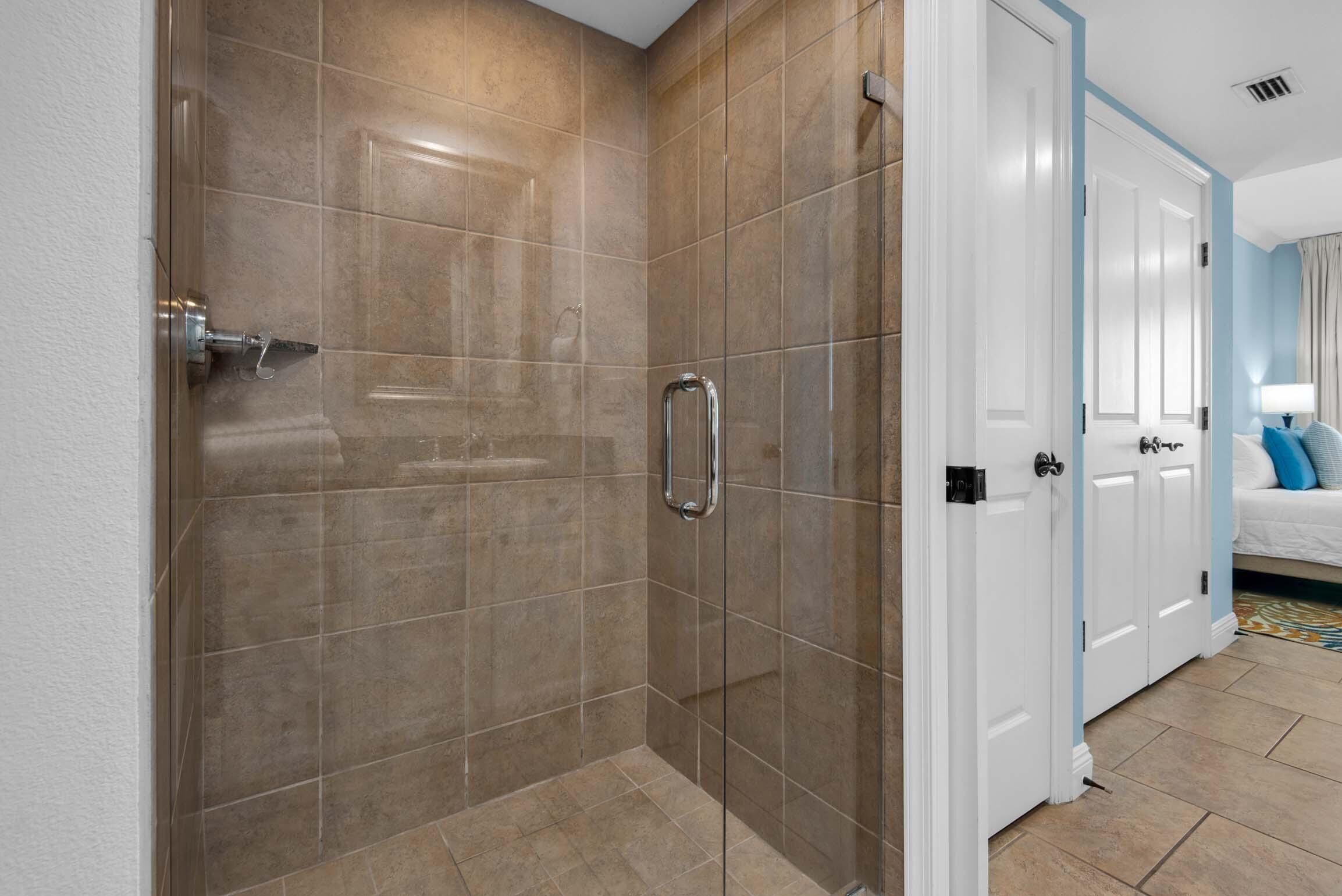 A modern bathroom with a glass shower enclosure, a walk-in shower, and a white door leading to a bedroom with a bed and a nightstand.