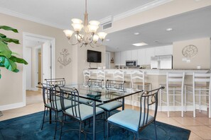 A dining area with a glass table, metal chairs, a blue rug, a chandelier, and a kitchen in the background. - Belle Mer 1102 (Navarre)