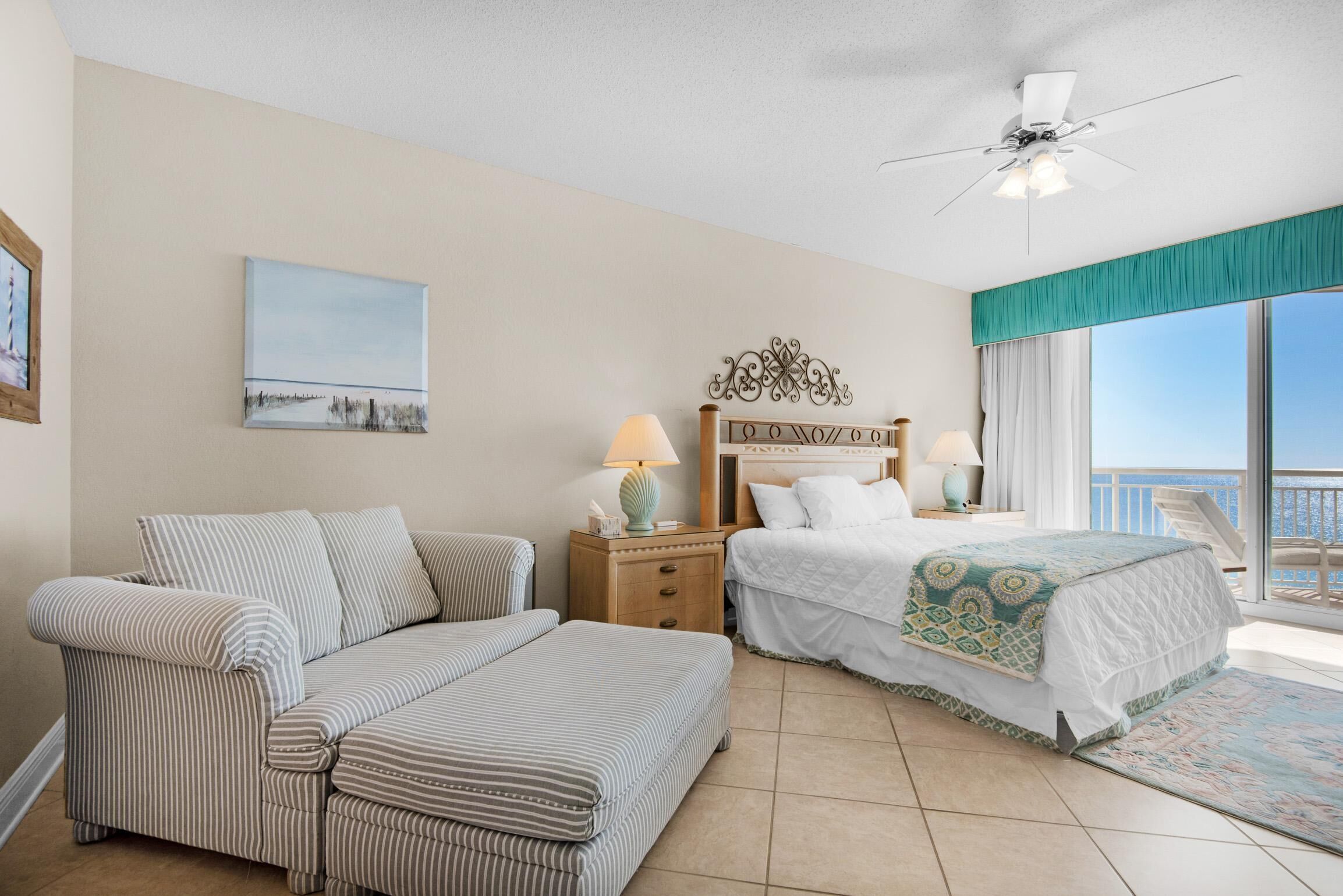 A hotel room with a bed, a sofa, a nightstand with lamps, a ceiling fan, a painting, and a view of the sea.