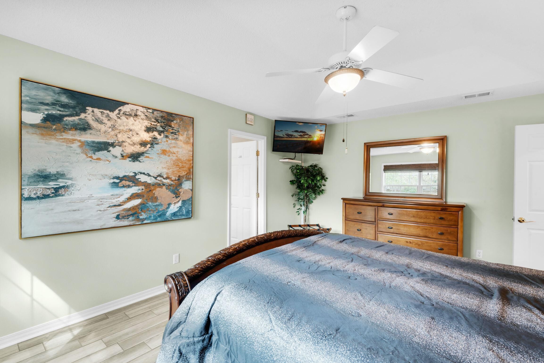 Une chambre à coucher comprenant un lit, un ventilateur de plafond, une télévision, une commode et un grand tableau accroché au mur.