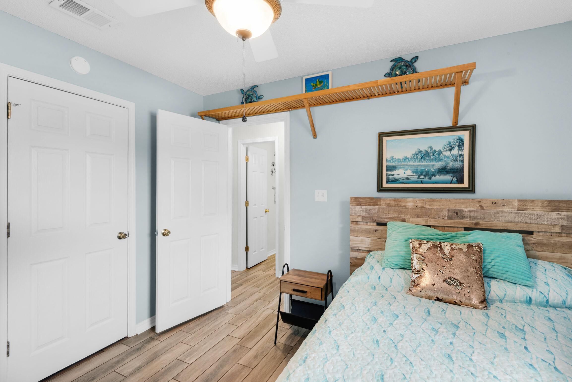 A bedroom with a wooden headboard, a bed with blue and white bedding, a small wooden nightstand, and a wall-mounted shelf with decorative items.