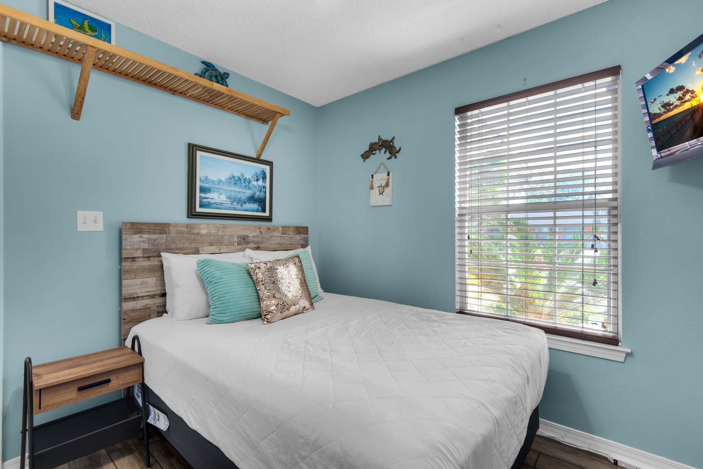 A bedroom with a bed, a window with blinds, a shelf with decorative items, and a framed picture on the wall.