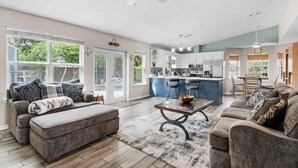 A spacious living room with a large window, a ceiling fan, and a kitchen area with bar stools. - Village Oasis (Gulf Breeze)