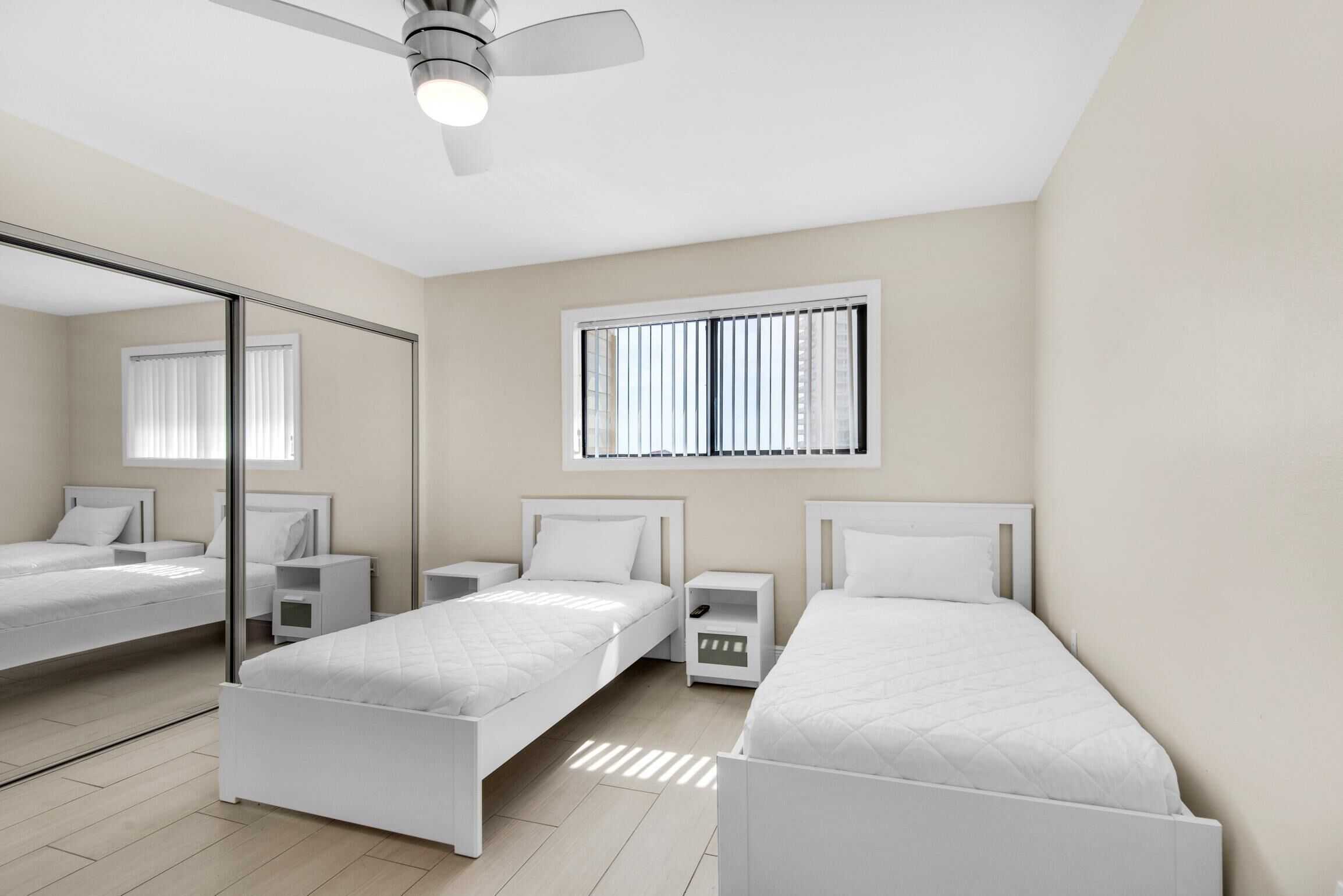 A modern bedroom with two single beds, a ceiling fan, a window with blinds, and a mirror on the wall.