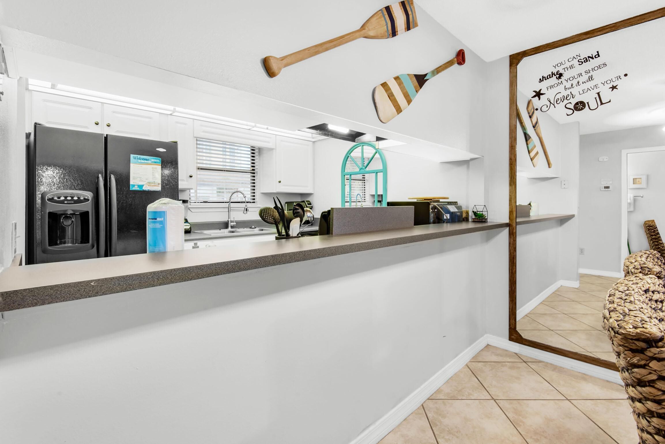 A kitchen with a refrigerator, a coffee machine, and a sink. There are wooden paddles hanging on the wall and a motivational quote.