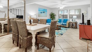 Uma área de refeições com mesa branca e cadeiras de vime, uma sala de estar com sofá azul e um espelho grande que reflete o espaço.