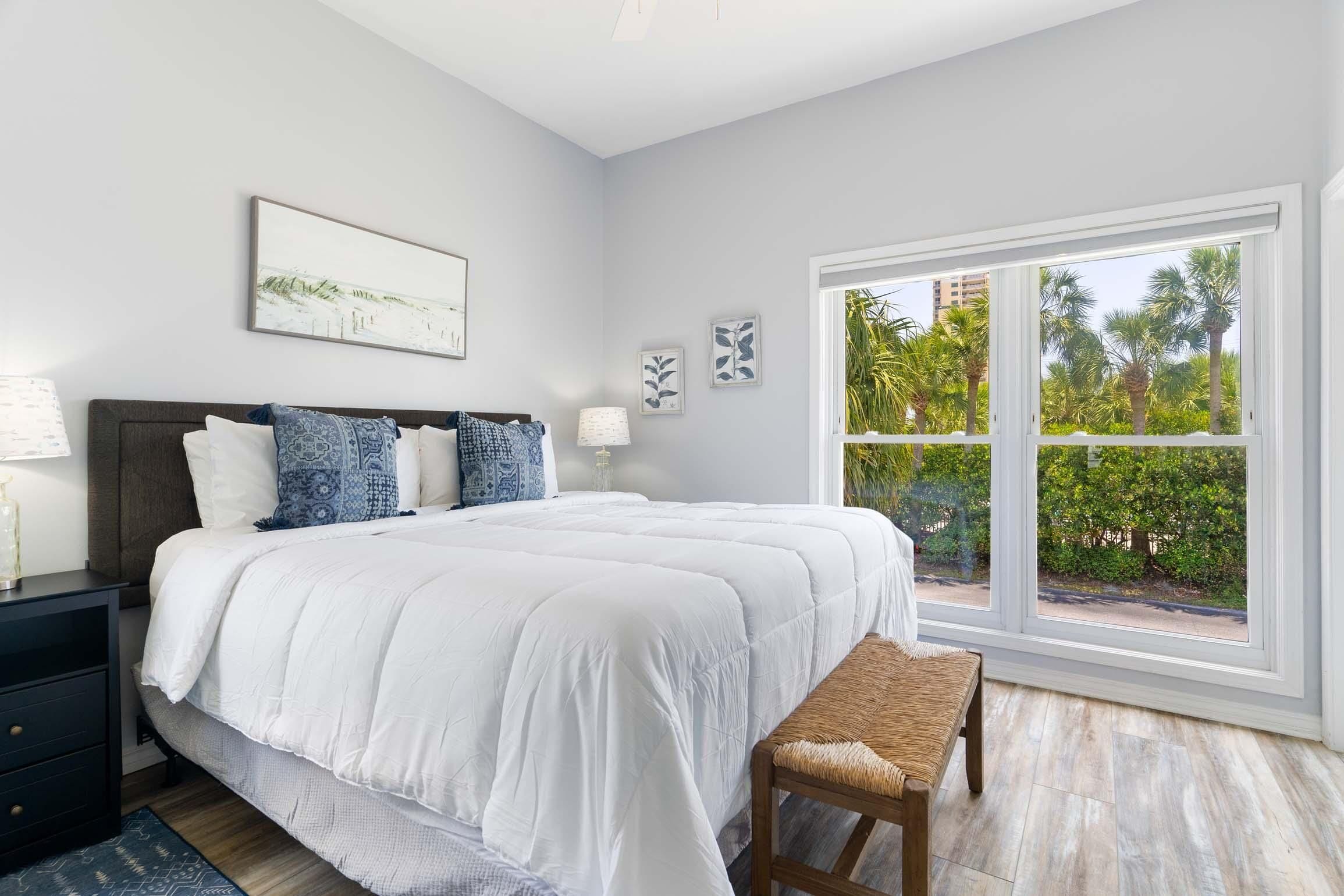 A bedroom with a large bed, a bedside table, a bench, and a window with a view of palm trees.